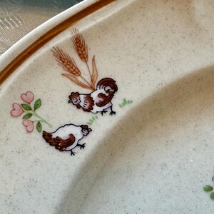 May include: A section of a cream-colored plate with a brown rim and speckled surface. The plate is decorated with illustrations of a rooster, a hen, wheat stalks, and floral accents in shades of brown, white, pink, and green.