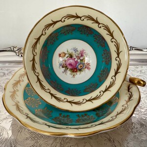 May include: A vintage teacup and saucer set. The teacup features a cream-colored rim with gold accents and a teal interior with a floral design. The saucer mirrors the cup's design, creating a cohesive and elegant set.