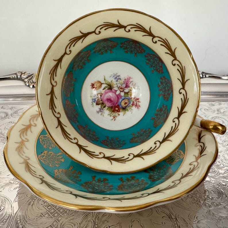 May include: A vintage teacup and saucer set. The teacup features a cream-colored rim with gold accents and a teal interior with a floral design. The saucer mirrors the cup's design, creating a cohesive and elegant set.