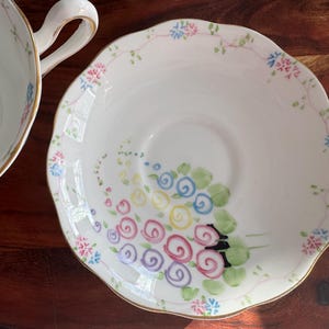May include: A white porcelain teacup and saucer with a floral design. The saucer has a scalloped edge and a gold trim. The floral design features pink, blue, yellow, and green flowers.