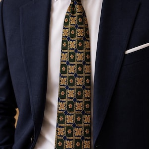 May include: A dark green necktie featuring a repeating grid pattern of gold floral motifs and small blue diamond shapes, worn with a crisp white dress shirt and a navy blue blazer with a white pocket square.