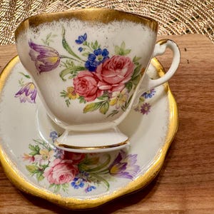 May include: A white teacup and saucer with a floral pattern in shades of pink, purple, and yellow. The cup and saucer have a gold trim.