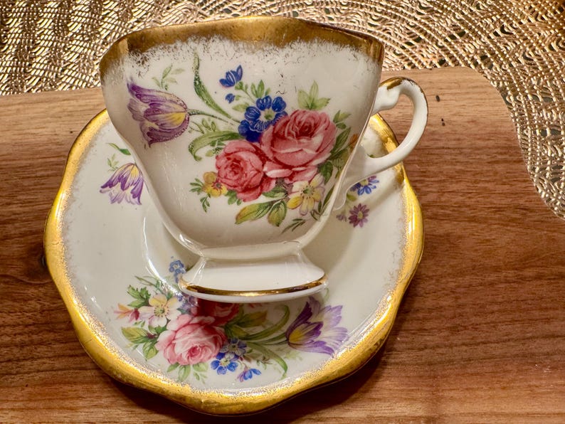 May include: A white teacup and saucer with a floral pattern in shades of pink, purple, and yellow. The cup and saucer have a gold trim.