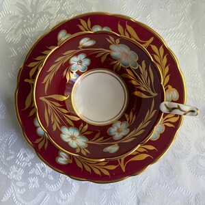 May include: A porcelain teacup and saucer set featuring a deep burgundy background with gold leaf foliage patterns and light blue floral accents. The teacup has a white interior base and a gold-trimmed handle with a delicate gold branch design.