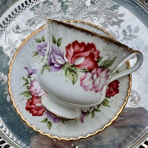 May include: A white teacup and saucer set with a floral design in red, pink, and purple. The set has gold trim and sits on a silver tray. The teacup has a curved handle.