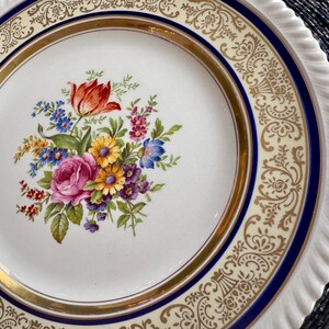 May include: A white ceramic plate with a floral bouquet design. The bouquet features a red tulip, pink rose, and other colorful flowers. The plate has a gold rim and a blue border with a decorative pattern.