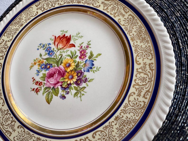 May include: A white ceramic plate with a floral bouquet design. The bouquet features a red tulip, pink rose, and other colorful flowers. The plate has a gold rim and a blue border with a decorative pattern.