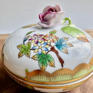 May include: A white porcelain trinket box with a floral design and a pink rose lid. The box has a gold trim and a green base.