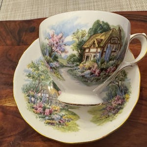 May include: A white teacup and saucer set with a cottage scene painted on both pieces. The scene features a small cottage with a thatched roof, a winding path, and lush greenery.