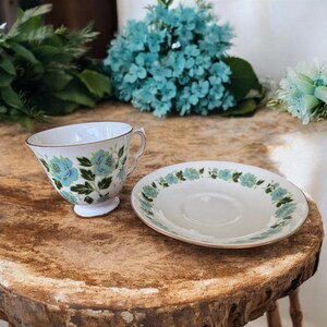 May include: A white teacup and saucer with a blue and green floral pattern. The cup and saucer have a gold trim.