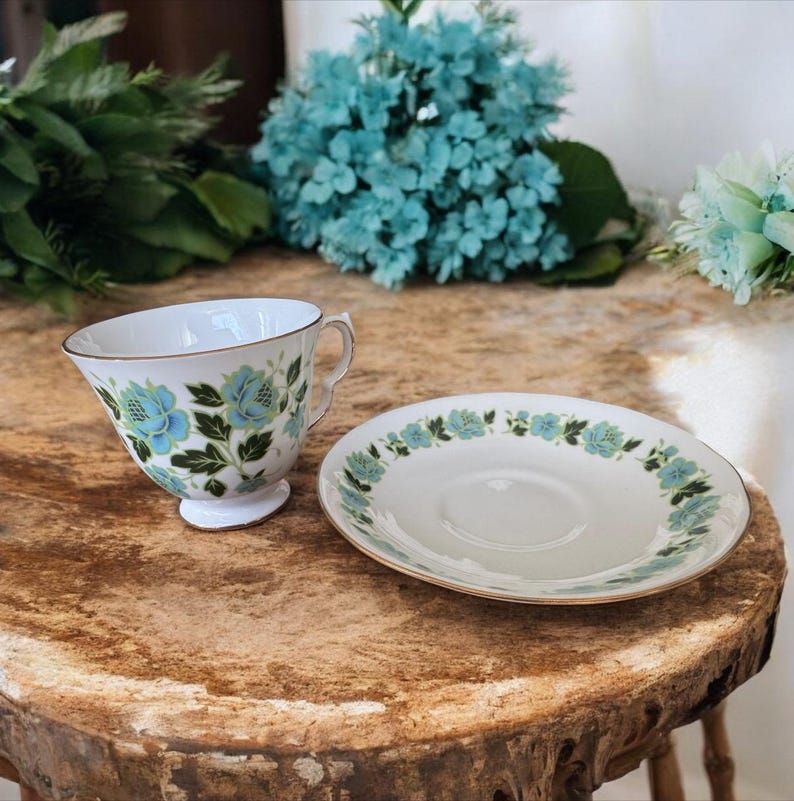 May include: A white teacup and saucer with a blue and green floral pattern. The cup and saucer have a gold trim.