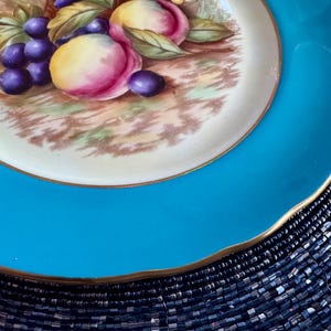 May include: A decorative plate with a vibrant turquoise rim and a gold edge. The center features a detailed painting of purple grapes and peaches with green leaves. The plate's design is classic and elegant, suitable for display or serving.