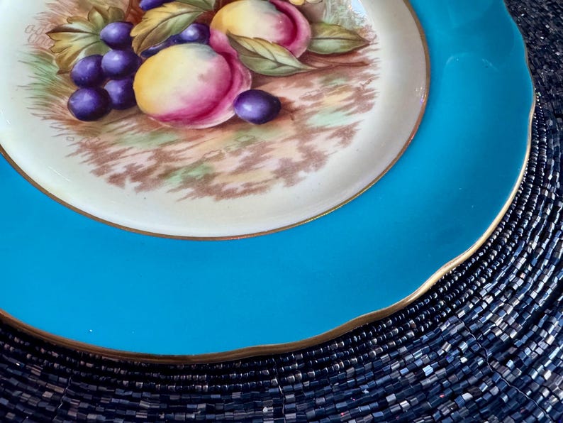 May include: A decorative plate with a vibrant turquoise rim and a gold edge. The center features a detailed painting of purple grapes and peaches with green leaves. The plate's design is classic and elegant, suitable for display or serving.