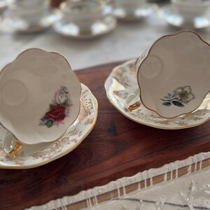 May include: Two white teacups with gold trim and floral designs on the side. The teacups are on matching saucers with gold trim and floral designs. The teacups and saucers are on a wooden surface.
