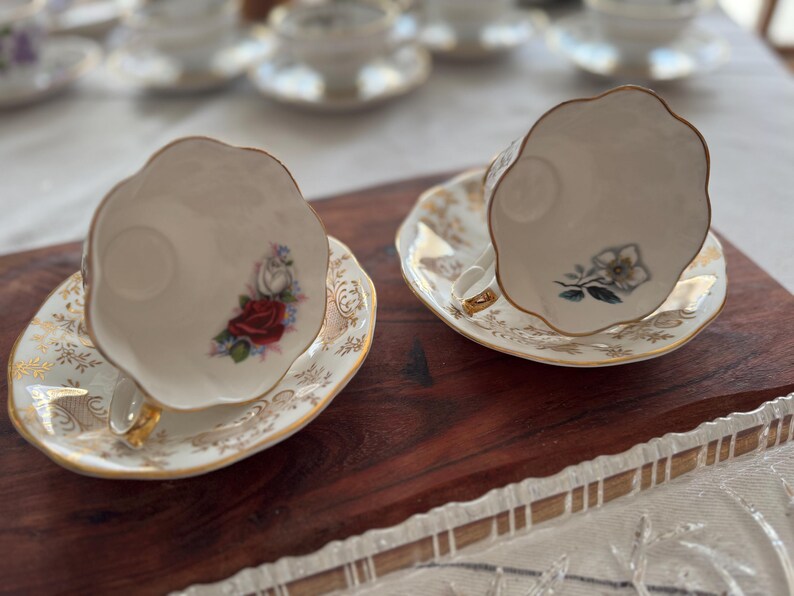 May include: Two white teacups with gold trim and floral designs on the side. The teacups are on matching saucers with gold trim and floral designs. The teacups and saucers are on a wooden surface.