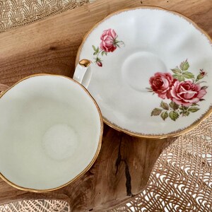 Pu&ograve; includere: Una tazza da t&egrave; e un piattino bianchi con un bordo dorato e un motivo floreale con rose rosa. Il piattino ha un bordo ondulato.