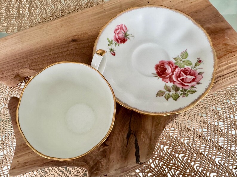 Pu&ograve; includere: Una tazza da t&egrave; e un piattino bianchi con un bordo dorato e un motivo floreale con rose rosa. Il piattino ha un bordo ondulato.