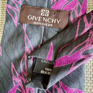 Puede incluir: Una corbata gris con un patr&oacute;n de hojas abstractas en rosa vibrante. Incluye una etiqueta marr&oacute;n con el texto GIVENCHY MONSIEUR y una etiqueta negra que indica 100% SILK y CA0008. La tela presenta una textura de tejido diagonal en toda ella.
