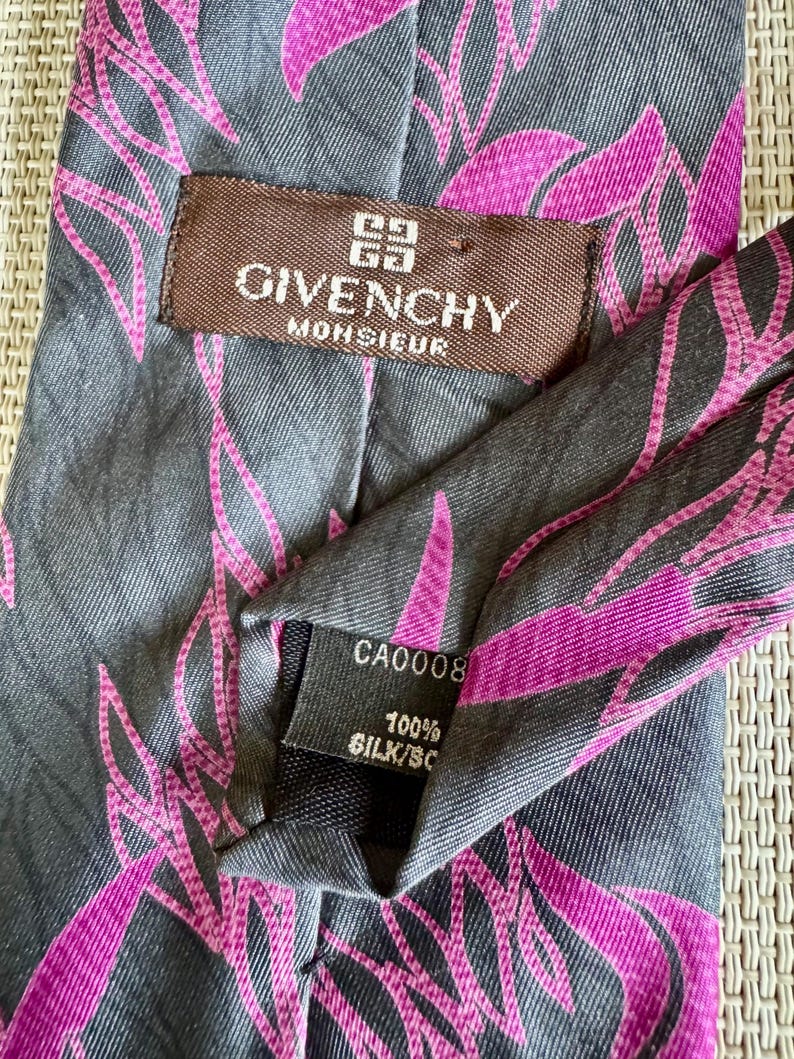 Puede incluir: Una corbata gris con un patr&oacute;n de hojas abstractas en rosa vibrante. Incluye una etiqueta marr&oacute;n con el texto GIVENCHY MONSIEUR y una etiqueta negra que indica 100% SILK y CA0008. La tela presenta una textura de tejido diagonal en toda ella.