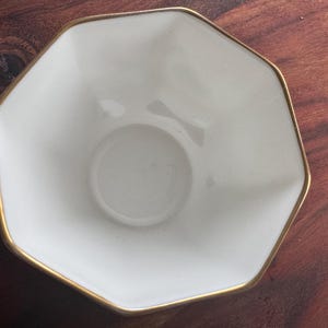Pode incluir: Uma tigela branca octogonal com borda dourada. A tigela tem uma pequena reentr&acirc;ncia circular no centro. A tigela est&aacute; sobre uma superf&iacute;cie de madeira marrom escura. Esta tigela &eacute; ideal para servir pequenas por&ccedil;&otilde;es de comida.