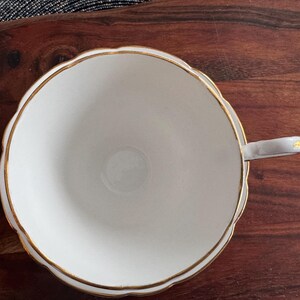 May include: A white teacup with a gold rim and a scalloped edge. The teacup has a handle and is sitting on a wooden surface.