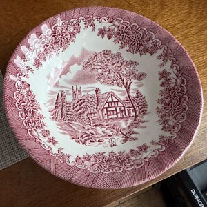 Pu&ograve; includere: Una ciotola rotonda in ceramica bianca e rossa con una scena paesaggistica dettagliata. La ciotola presenta un bordo floreale rosso e un'immagine centrale di una casa, alberi e una chiesa. La ciotola &egrave; probabilmente vintage o antica.