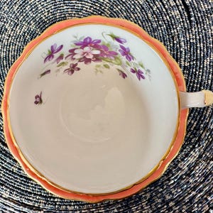 May include: A white teacup with a pink rim and gold trim. The cup is decorated with a floral pattern of purple and pink flowers.