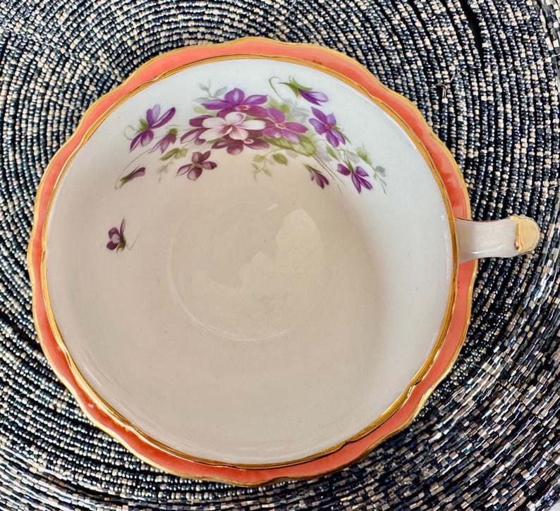 May include: A white teacup with a pink rim and gold trim. The cup is decorated with a floral pattern of purple and pink flowers.