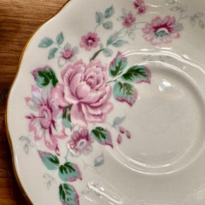 May include: A white saucer with a gold rim features a floral design with pink and green roses.