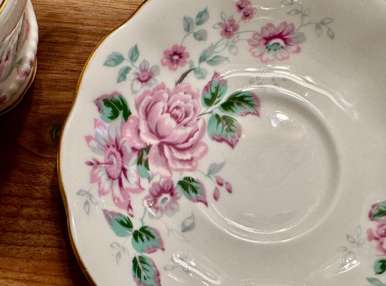 May include: A white saucer with a gold rim features a floral design with pink and green roses.