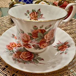 Pode incluir: Uma ch&aacute;vena de ch&aacute; e um pires brancos com uma borda dourada e um padr&atilde;o floral de rosas cor-de-rosa e folhas verdes.