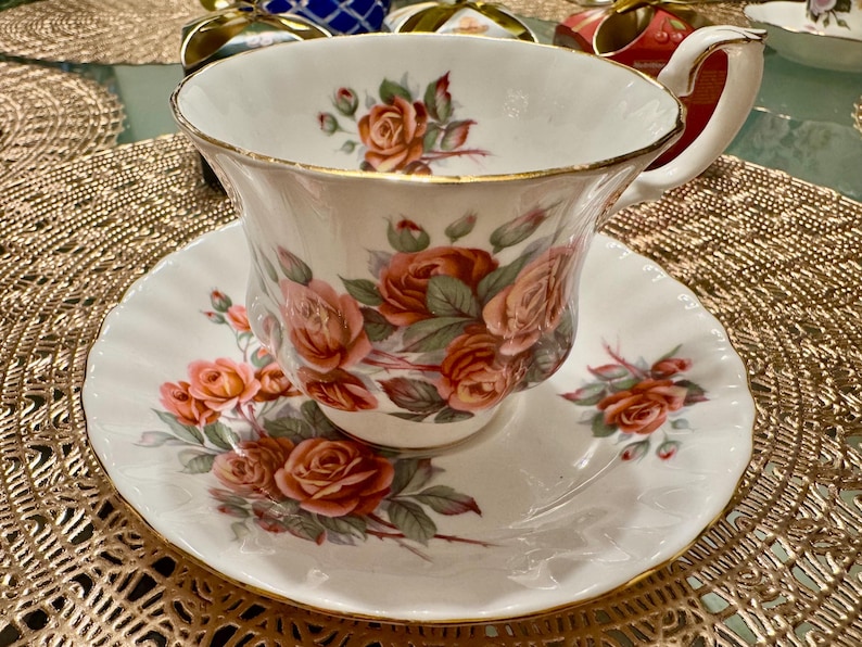 Pode incluir: Uma ch&aacute;vena de ch&aacute; e um pires brancos com uma borda dourada e um padr&atilde;o floral de rosas cor-de-rosa e folhas verdes.