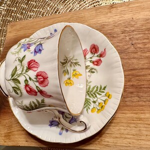May include: A white teacup and saucer with a floral pattern. The pattern features pink, red, blue, yellow, and green flowers. The cup and saucer have a gold trim.