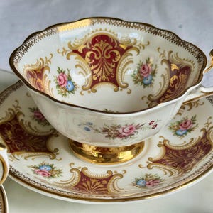 May include: A vintage teacup and saucer set. The white porcelain cup and saucer feature a burgundy and gold ornate design with floral accents. The cup has a gold handle and trim, and the saucer has a matching border.