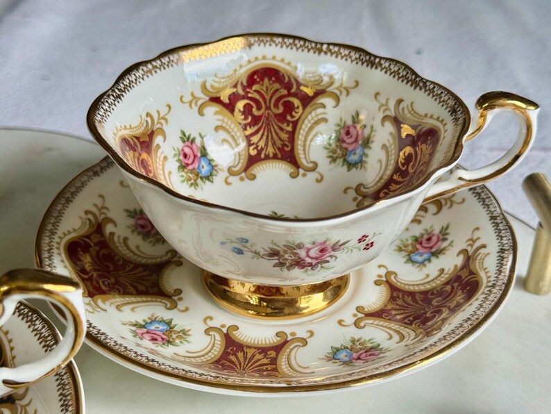 May include: A vintage teacup and saucer set. The white porcelain cup and saucer feature a burgundy and gold ornate design with floral accents. The cup has a gold handle and trim, and the saucer has a matching border.
