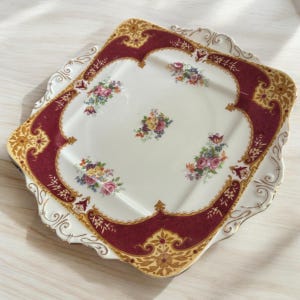 May include: A square ceramic serving plate with a scalloped edge. The plate has a white background with a red and gold floral design.
