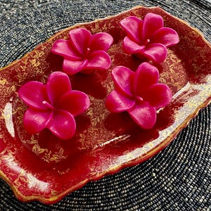 May include: Four pink flower-shaped candles on a red and gold decorative tray. The tray has a scalloped edge and a floral design.