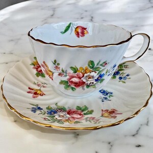 Puede incluir: Una taza de t&eacute; y un platillo blancos con un patr&oacute;n floral. La taza y el platillo tienen un borde dorado. Las flores son rosas, amarillas y azules.
