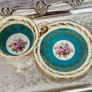 May include: A vintage teacup and saucer set. The cup and saucer feature a teal and cream color scheme with gold accents. Floral designs are present in the center of the cup and saucer. The set is placed on a silver tray.