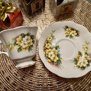 May include: A white teacup and saucer with a floral design featuring yellow primrose flowers. The cup and saucer are decorated with a delicate floral pattern.
