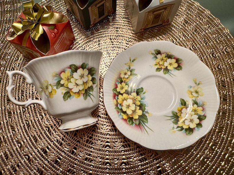 May include: A white teacup and saucer with a floral design featuring yellow primrose flowers. The cup and saucer are decorated with a delicate floral pattern.