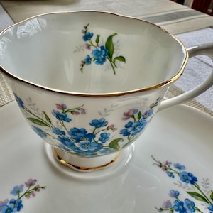 May include: A white teacup and saucer set with a blue and white floral pattern. The cup has a gold rim and the saucer has a floral design around the edge.