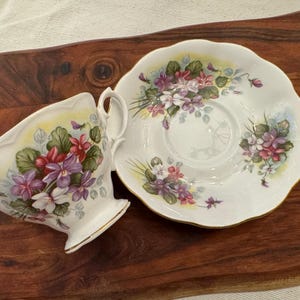 May include: A white teacup and saucer set with a floral design featuring purple, pink, and green violets. The set has a gold trim around the edges.