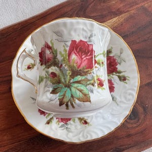 May include: A white teacup and saucer with a floral design featuring red roses and green leaves. The cup has a gold rim and a delicate handle.