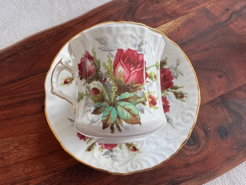 May include: A white teacup and saucer with a floral design featuring red roses and green leaves. The cup has a gold rim and a delicate handle.