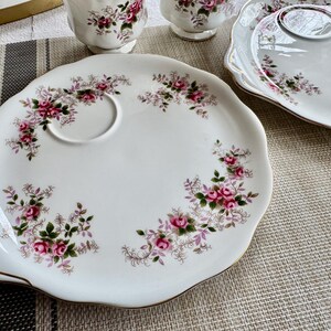 May include: A white porcelain tea plate with a floral design featuring pink roses and green leaves. The plate has a scalloped edge and a gold rim. There is a smaller, circular indentation in the plate for a cup.