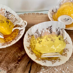 Puede incluir: Cuatro tazas y platillos vintage. Las tazas son amarillas con bordes blancos y detalles florales dorados. Los platillos tienen dise&ntilde;os florales dorados a juego. Las tazas y platillos est&aacute;n colocados sobre una superficie de madera.