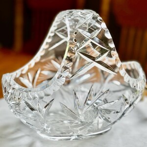 May include: A clear, cut-glass basket with a handle. The basket features a starburst pattern on the base and sides, with diamond and geometric cuts throughout. The handle is also cut with a diamond pattern. The basket sits on a white marble surface.