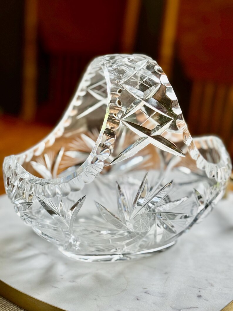 May include: A clear, cut-glass basket with a handle. The basket features a starburst pattern on the base and sides, with diamond and geometric cuts throughout. The handle is also cut with a diamond pattern. The basket sits on a white marble surface.