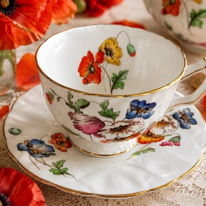 K&ouml;nnte beinhalten: Wei&szlig;es Tee-Set mit Blumendesign aus roten, gelben und blauen Blumen. Die Tasse und Untertasse haben einen goldenen Rand. Rote Mohnblumen sind im Hintergrund.